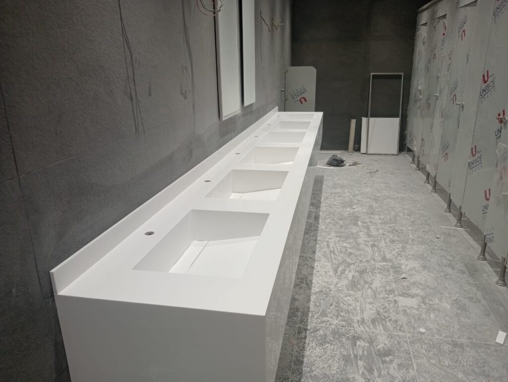 Seamless white Corian solid surface vanity trough with integrated moulded basins installed by Cairo House in a commercial washroom in Cairo, compact HPL partitions behind, showing one continuous surface with no join between the counter and the sinks.