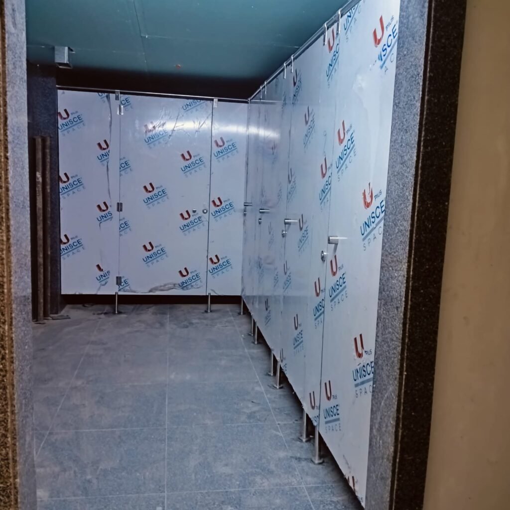 Compact HPL partitions in Cairo down a narrow washroom corridor, panels still under blue UNISCE film. Cairo House works to tight commercial washroom layouts with compact HPL bathroom partitions across Cairo.