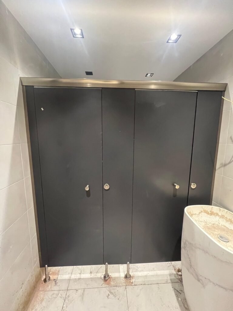 Three charcoal compact HPL cubicles with stainless rail and legs in a white tiled washroom, an onyx pedestal basin in front. One of the compact HPL in Cairo projects by Cairo House (كايرو هاوس - قواطيع حمامات كوريان و كومباكت HPL و ارضيات مرتفعة).