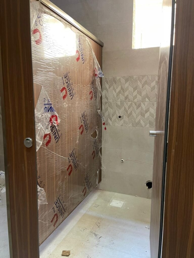 Dark walnut wood grain compact HPL cubicle door, still half covered in its UNISCE protective film, framed by a timber doorway and grey chevron tiles. Compact HPL in Cairo by Cairo House (كايرو هاوس - قواطيع حمامات كوريان و كومباكت HPL و ارضيات مرتفعة).