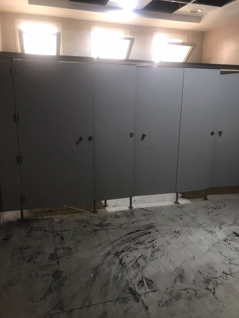 Grey compact HPL cubicle bank under bright skylights, doors closed, stainless legs on a white marble floor before final cleaning. A compact HPL in Cairo washroom fitted by Cairo House (كايرو هاوس - قواطيع حمامات كوريان و كومباكت HPL و ارضيات مرتفعة).