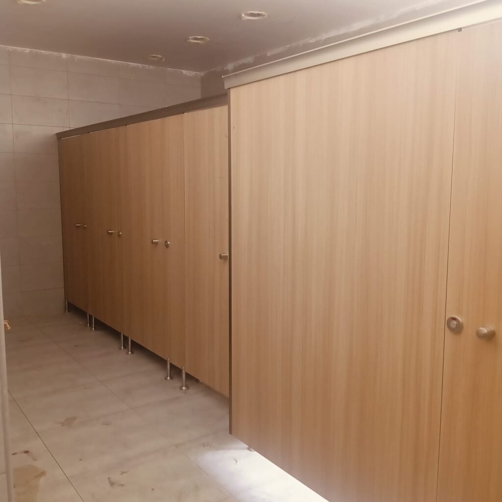 A long run of light oak compact HPL toilet cubicles in warm light, steel legs on a cream tiled floor and one door ajar at the end. Compact HPL in Cairo installed by Cairo House (كايرو هاوس - قواطيع حمامات كوريان و كومباكت HPL و ارضيات مرتفعة).