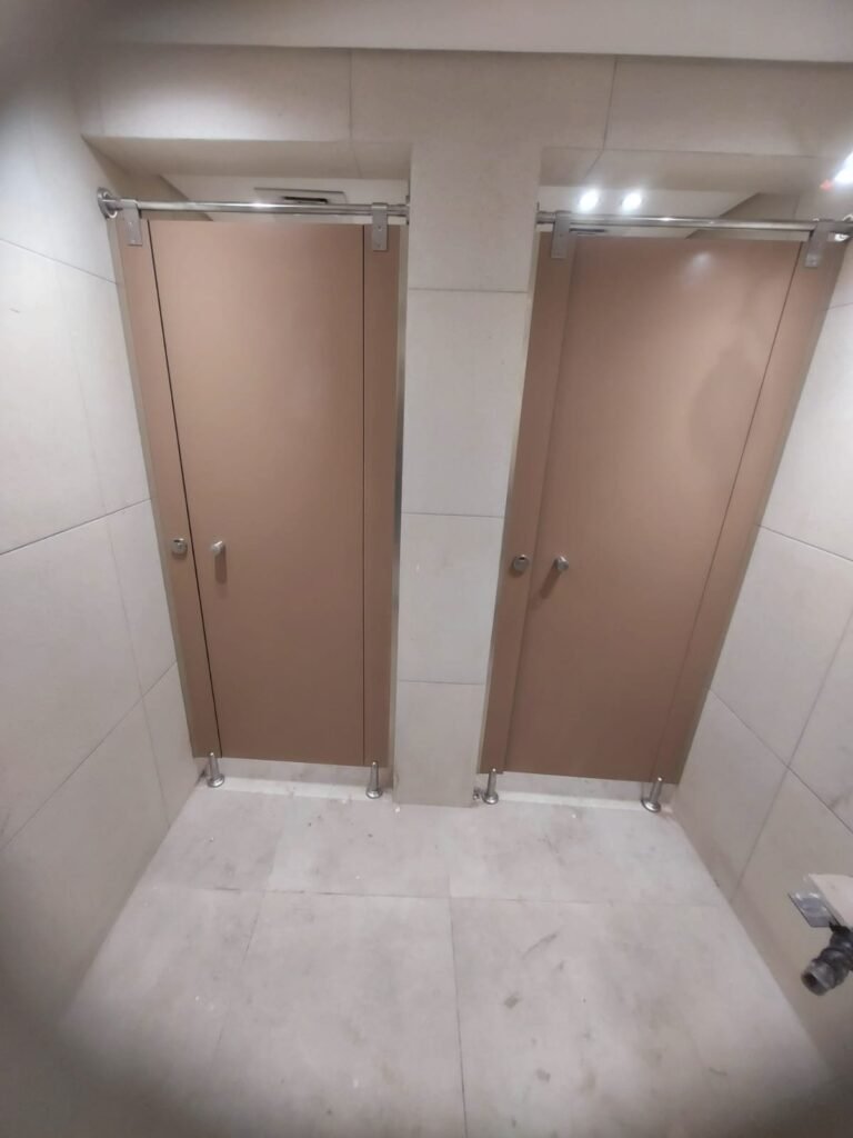 Two mocha brown compact HPL toilet doors side by side between beige wall tiles, with steel headrail and round knob locks. A finished compact HPL in Cairo washroom from Cairo House (كايرو هاوس - قواطيع حمامات كوريان و كومباكت HPL و ارضيات مرتفعة).